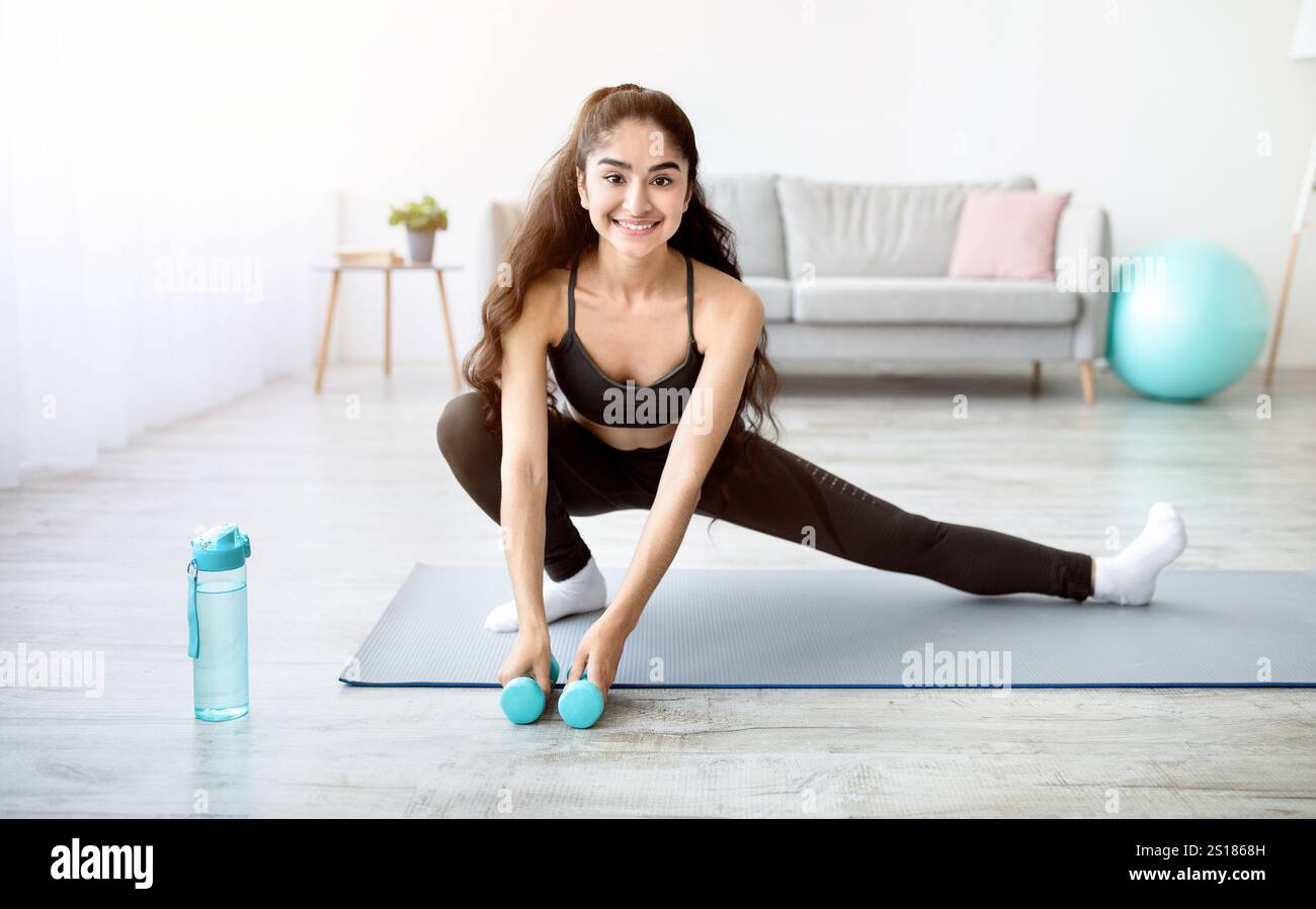 Young Indian woman doing stretching exercises with dumbbells on mat in ...