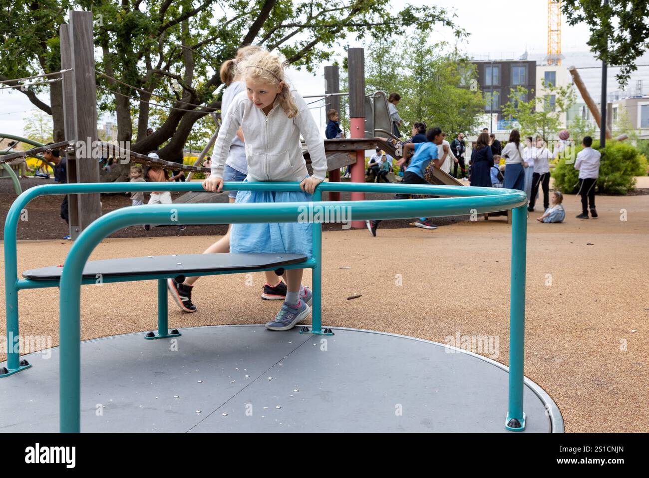 A 7 year old white girl spins on a roundabout in a playground at Brent Cross Town, a development in NW London. Her 8 year old sister is behind her. Stock Photo