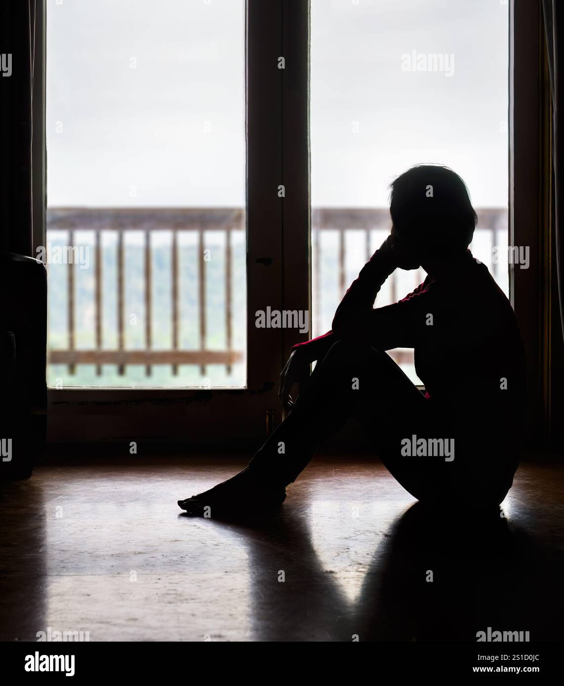 Lonely woman sitting on the floor by the glass door, looking outside. Stock Photo