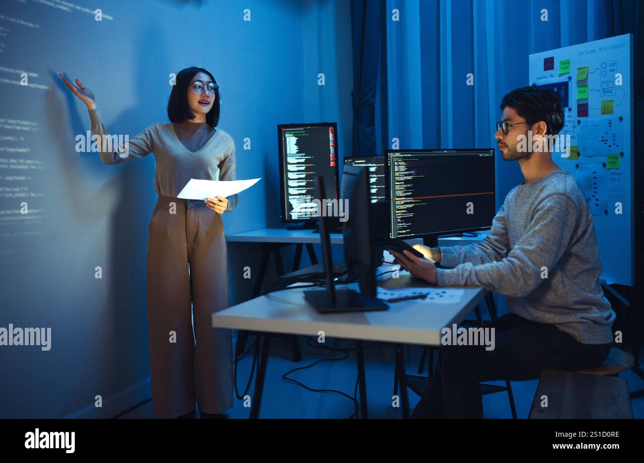 asian woman developers collaborating on a coding project, with one member presentation at code projected on the wall while the other works on multiple Stock Photo