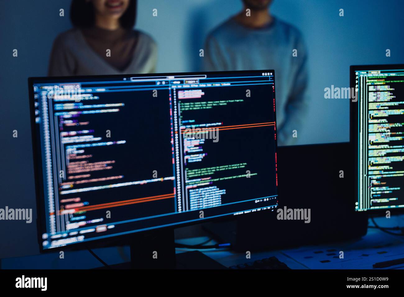Close-up of computer screens displaying code, with developers collaborating in the background Stock Photo