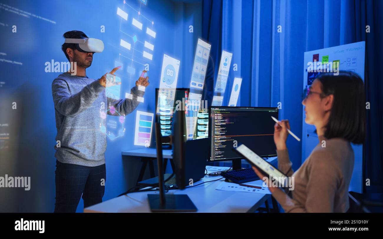 developer uses a VR headset to interact with projected interface designs, while colleagues discuss and code on screens at office Stock Photo