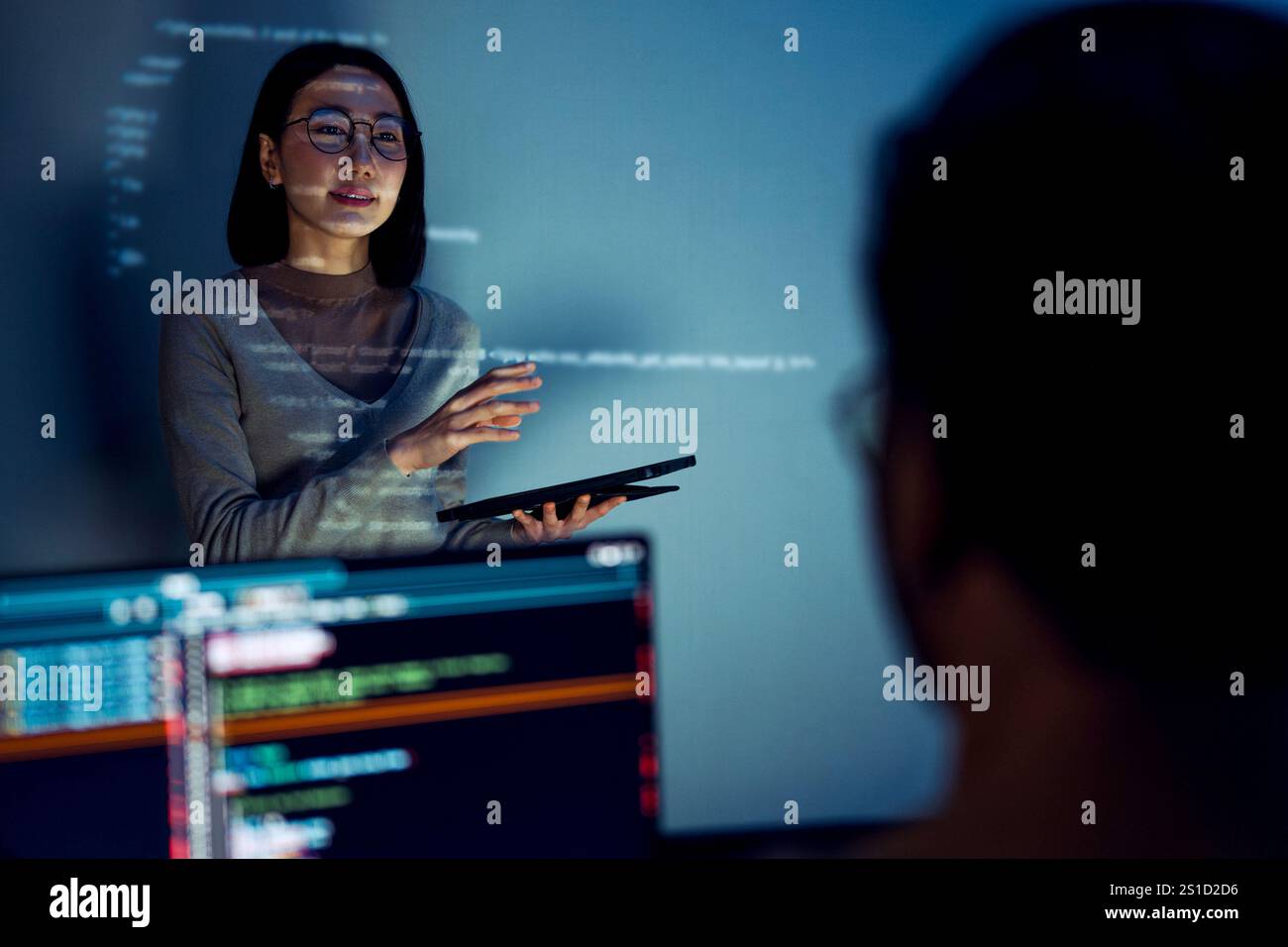 asian woman developers collaborating on a coding project, with one member presentation at code projected on the wall while the other works on multiple Stock Photo