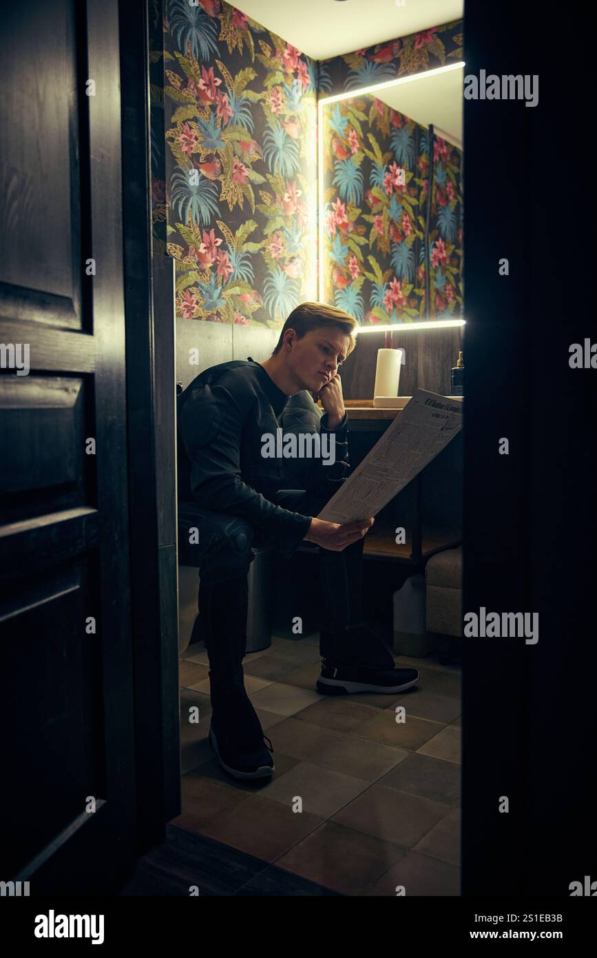 Man, dressed as superman, reading newspaper in dim bathroom, thoughtful ...