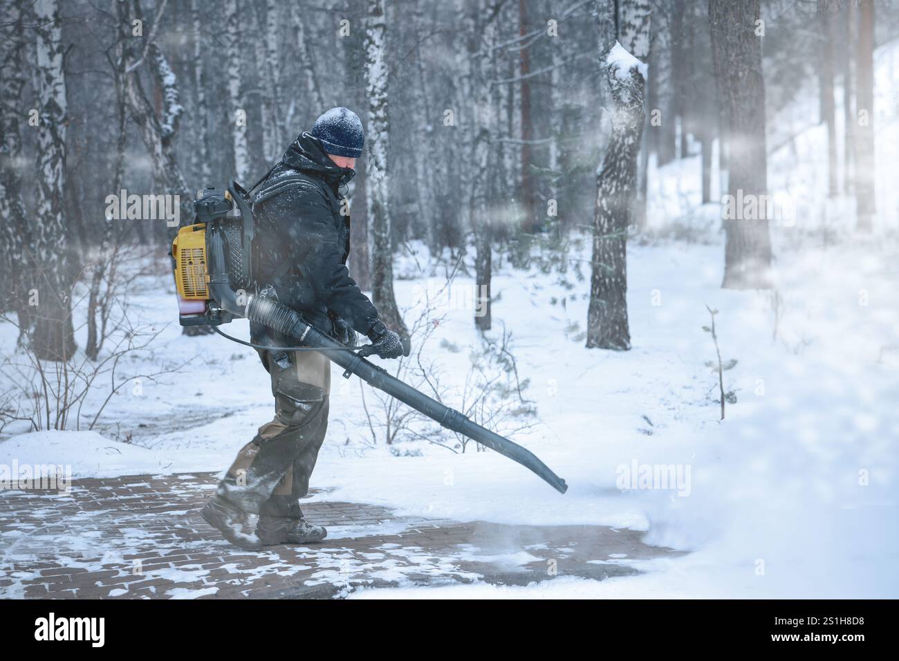 Worker using a leaf blower to clear snow in a winter forest, dressed in ...