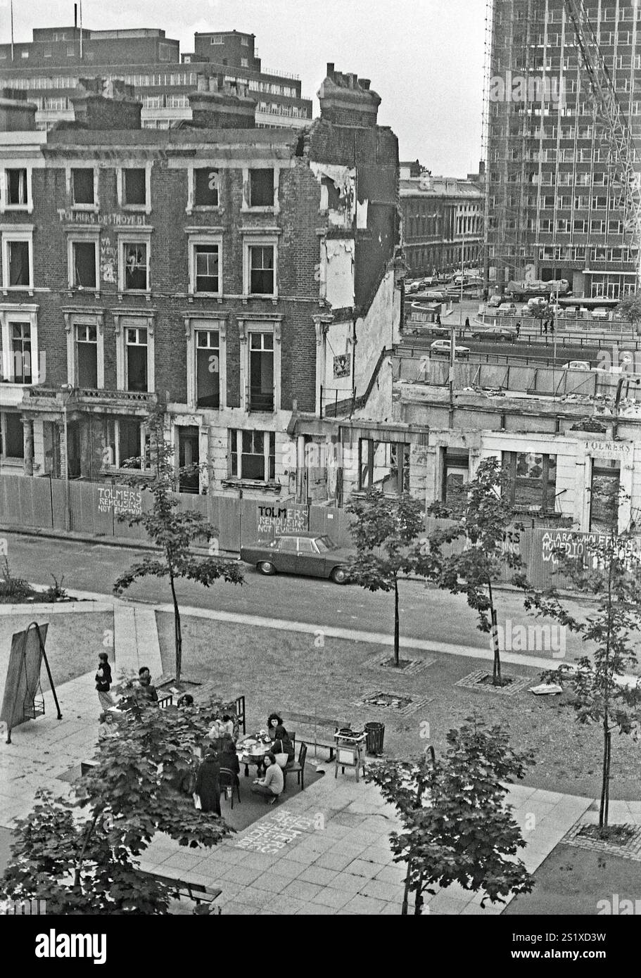 A view over Tolmers Square, Somers Town, London, England, UK, during the protests there in the late 1970s. Local residents are sitting in the square and hand-painted slogans can be seen on the buildings, some nearly demolished, and corrugated-iron fencing. Located in Somers Town, near Euston station in central London, Tolmers Square was occupied by more than one hundred squatters in the 1970s, who, along with local groups, fought for a redevelopment plan which fitted the local community. By 1975 ‘Tolmers Village’ had 49 squats housing over 180 people. The squatters lived there for six years. Stock Photo