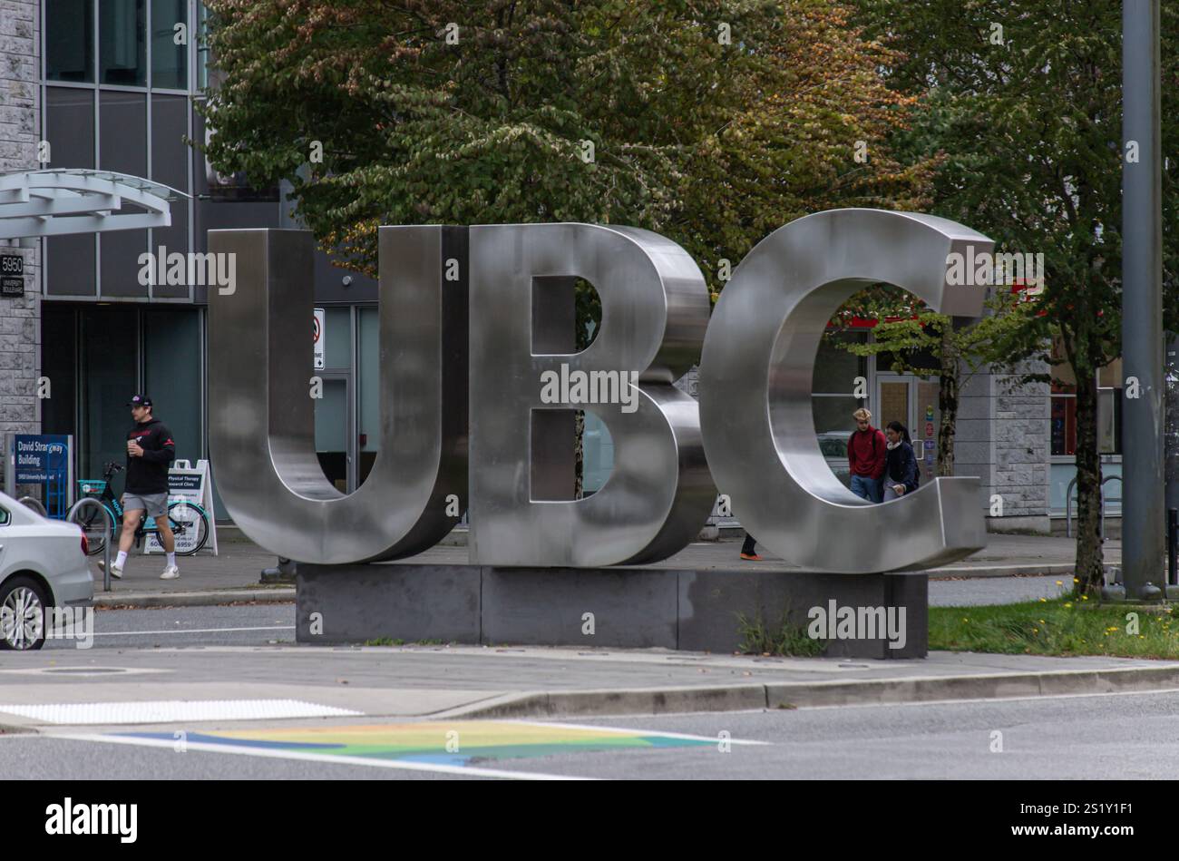 Large metallic UBC (The University of British Columbia) sign on a university campus surrounded by trees, pedestrians, and modern buildings. Stock Photo