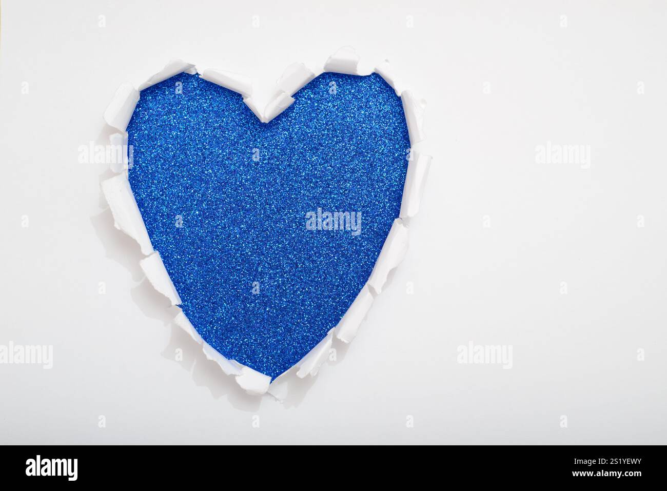 Heart-shaped hole torn on white paper revealing a blue glitter background. Copy space. Stock Photo