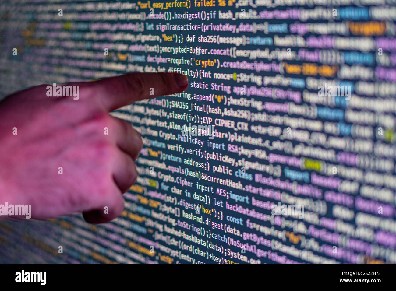 Close-up of a pointing hand over a large, glowing display screen filled with colorful code, possibly representing computer programming or data visuali Stock Photo