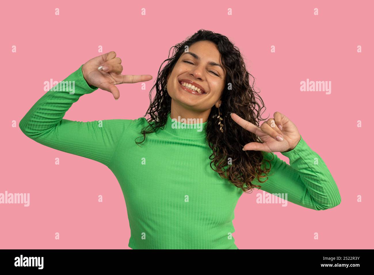 Cheerful young woman pointing herself with both index fingers, smiling and wearing a green turtleneck shirt on a vibrant pink background Stock Photo