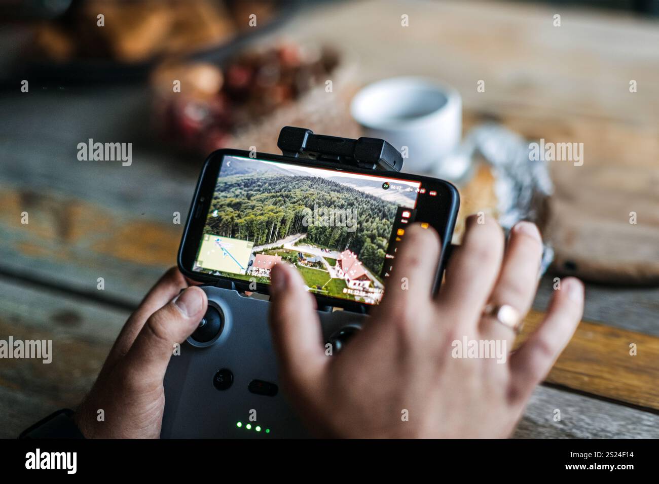 Close-up of hands using a smartphone and drone controller to navigate and view live aerial footage of a scenic landscape with forest and houses. Stock Photo