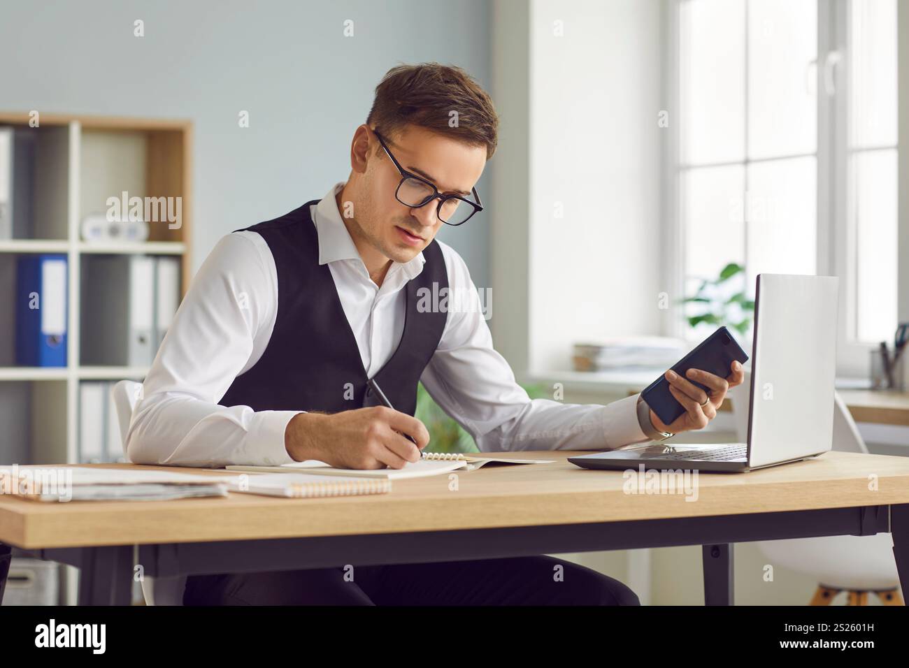 Serious businessman sitting at office desk with laptop, holding smartphone and writing in notebook Stock Photo