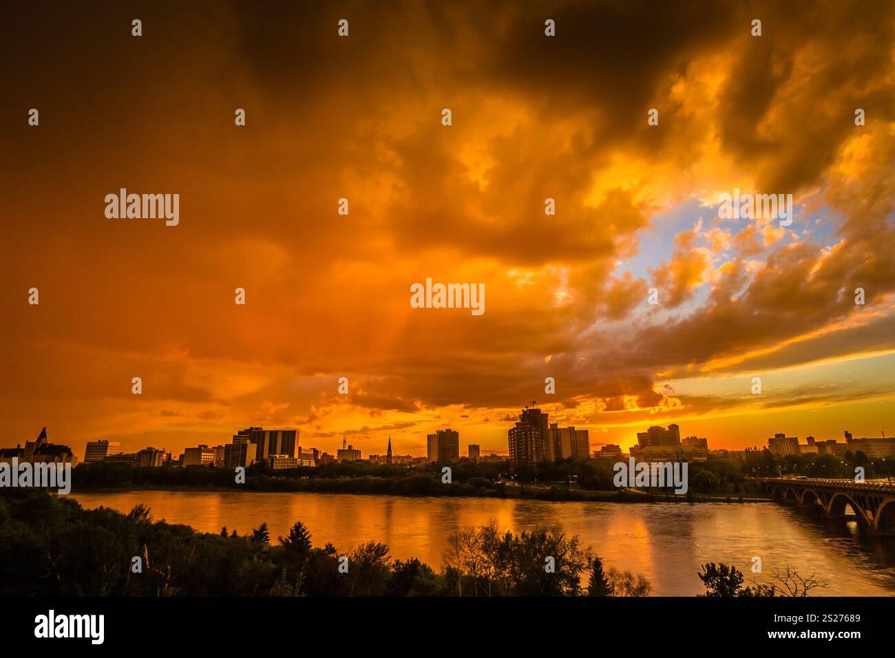 A city skyline is reflected in the water, with a beautiful orange sunset in the background. The sky is cloudy, but the sun is still visible, creating Stock Photo