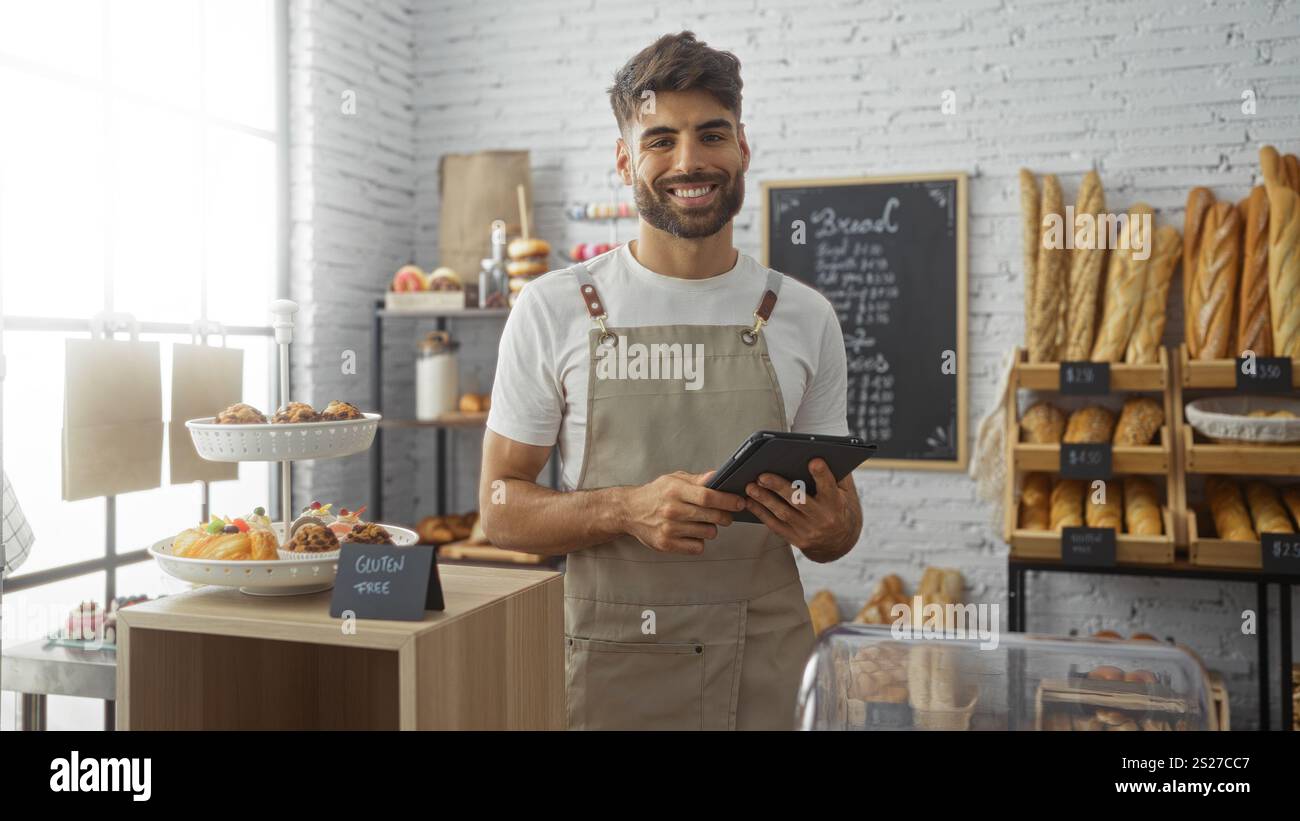 Young man with a beard working in a bakery shop, holding a tablet, surrounded by various types of bread and pastries displayed on shelves and counters Stock Photo