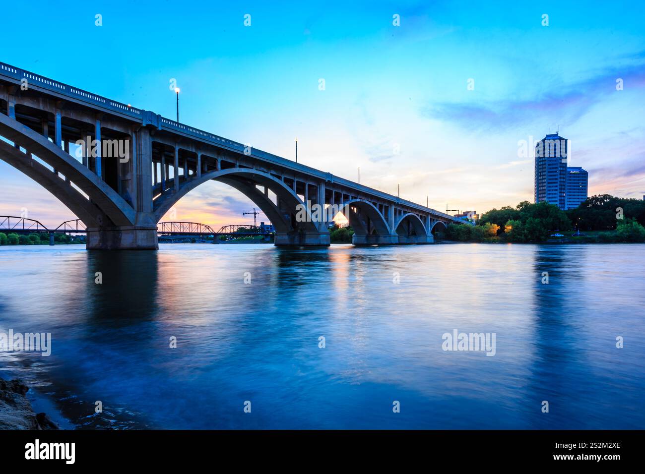 A bridge spans a river with a city skyline in the background. The sky is a beautiful blue color with a pinkish hue Stock Photo