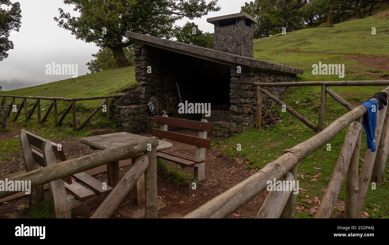 A rustic stone shelter with a wooden picnic table and benches in a green countryside setting. The fenced area and chimney suggest a barbecue or rest a Stock Photo