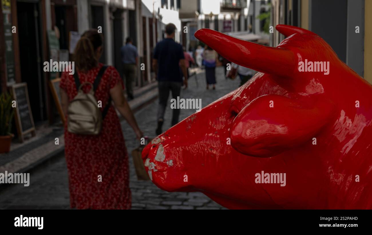 A red bull sculpture on a cobblestone street with pedestrians walking ...