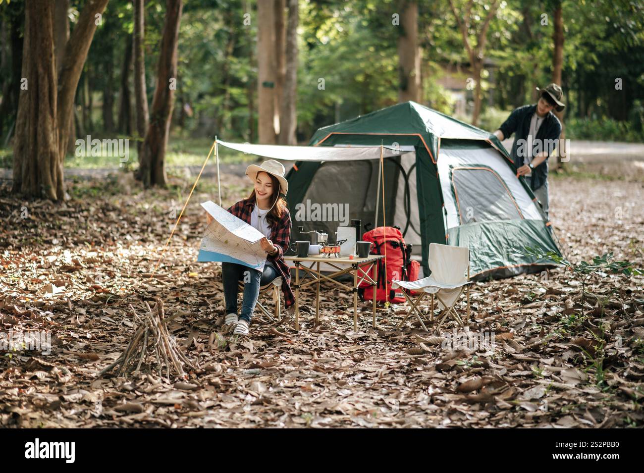 Pretty woman sitting on chair at front of camping tent and checking direction on paper map, Handsome boyfriend is pitching a tent behind her, They are Stock Photo