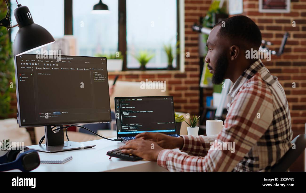 Young man seen programming software code on his laptop in a sleek home office. Workspace with dual monitors helps with software engineering, web development and coding productivity. Camera B. Stock Photo