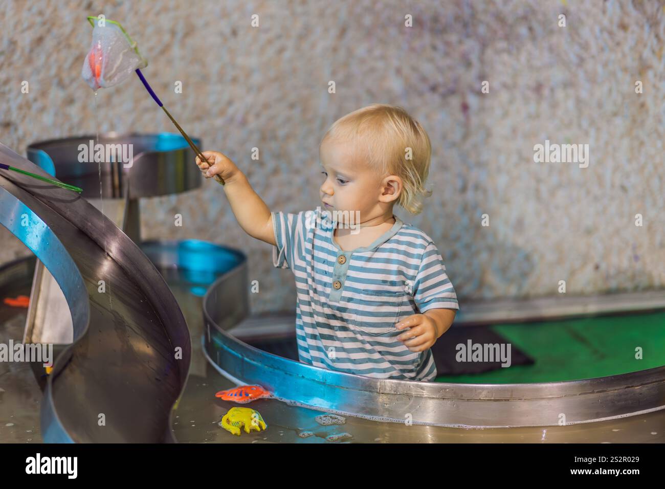 Toddler boy catching toy fish with a net in a vibrant water play area ...