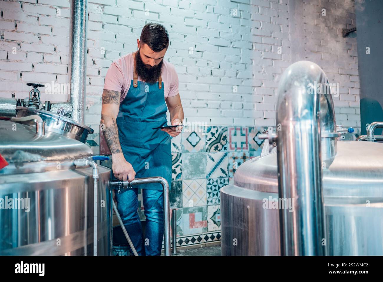 Bearded handsome man operating brewing equipment at beer making factory and using digital tablet. Beer production process, brewing at brewery. Stock Photo