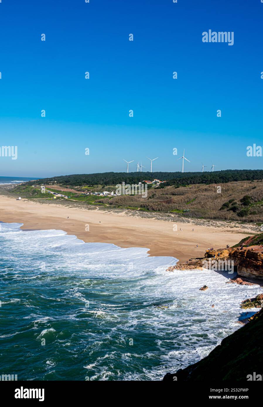 Nazaré, Portugal - Crashing waves and cliffs frame wind turbines on distant hills, combining natural beauty and renewable energy. Stock Photo