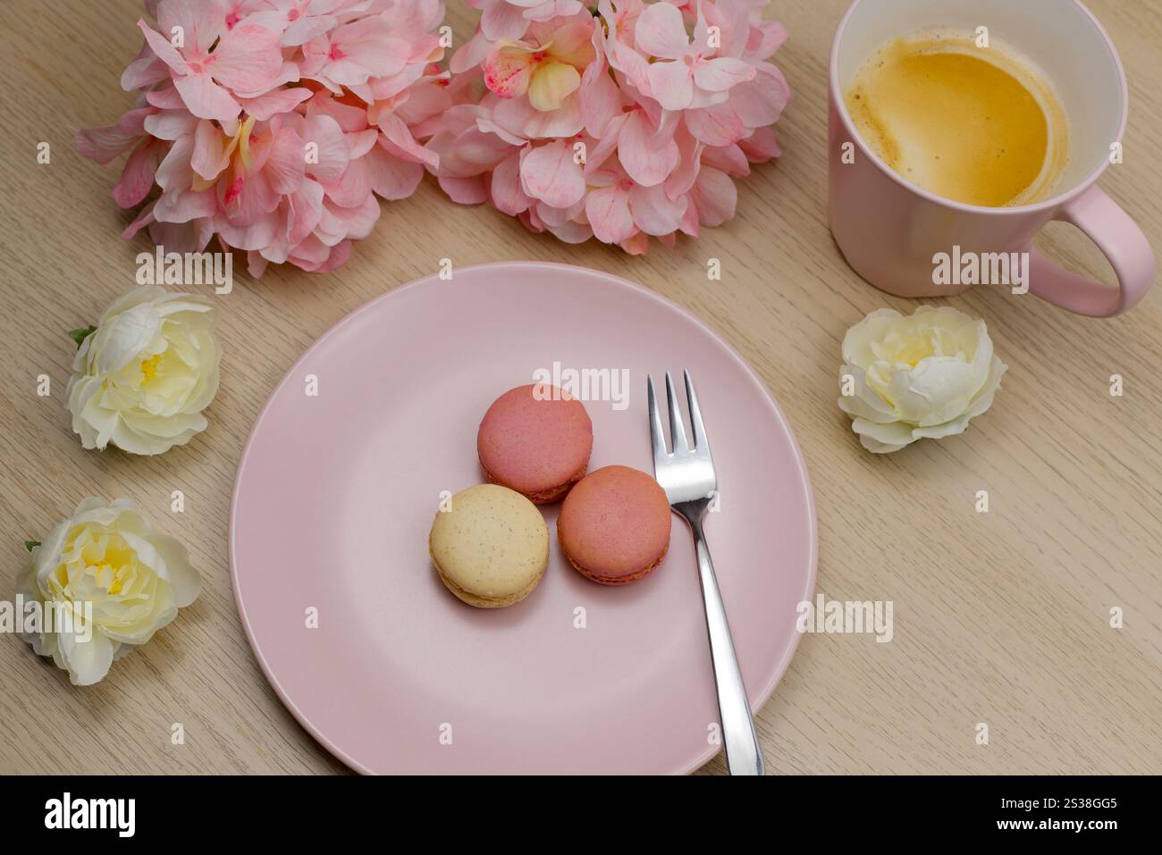 A mug of coffee with French raspberry and vanilla macarons on a wooden table with pink and cream coloured flowers. Romantic background Stock Photo