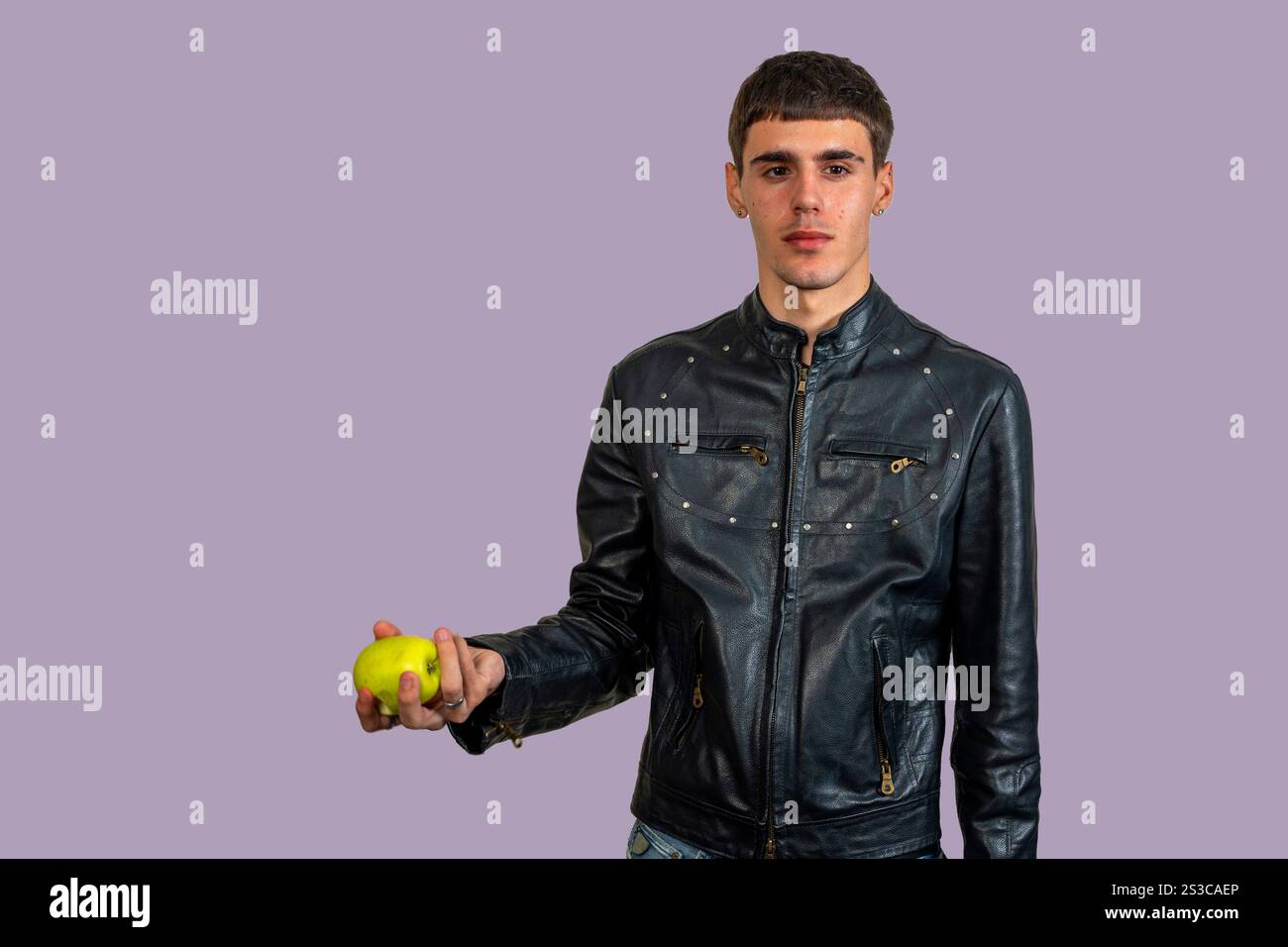 Young man wearing black leather jacket offering a green apple on neutral background Stock Photo