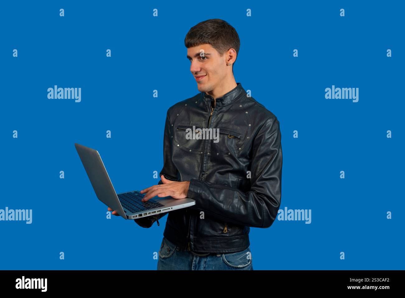 Young programmer with black leather jacket is using a laptop and smiling while looking away on a blue background Stock Photo