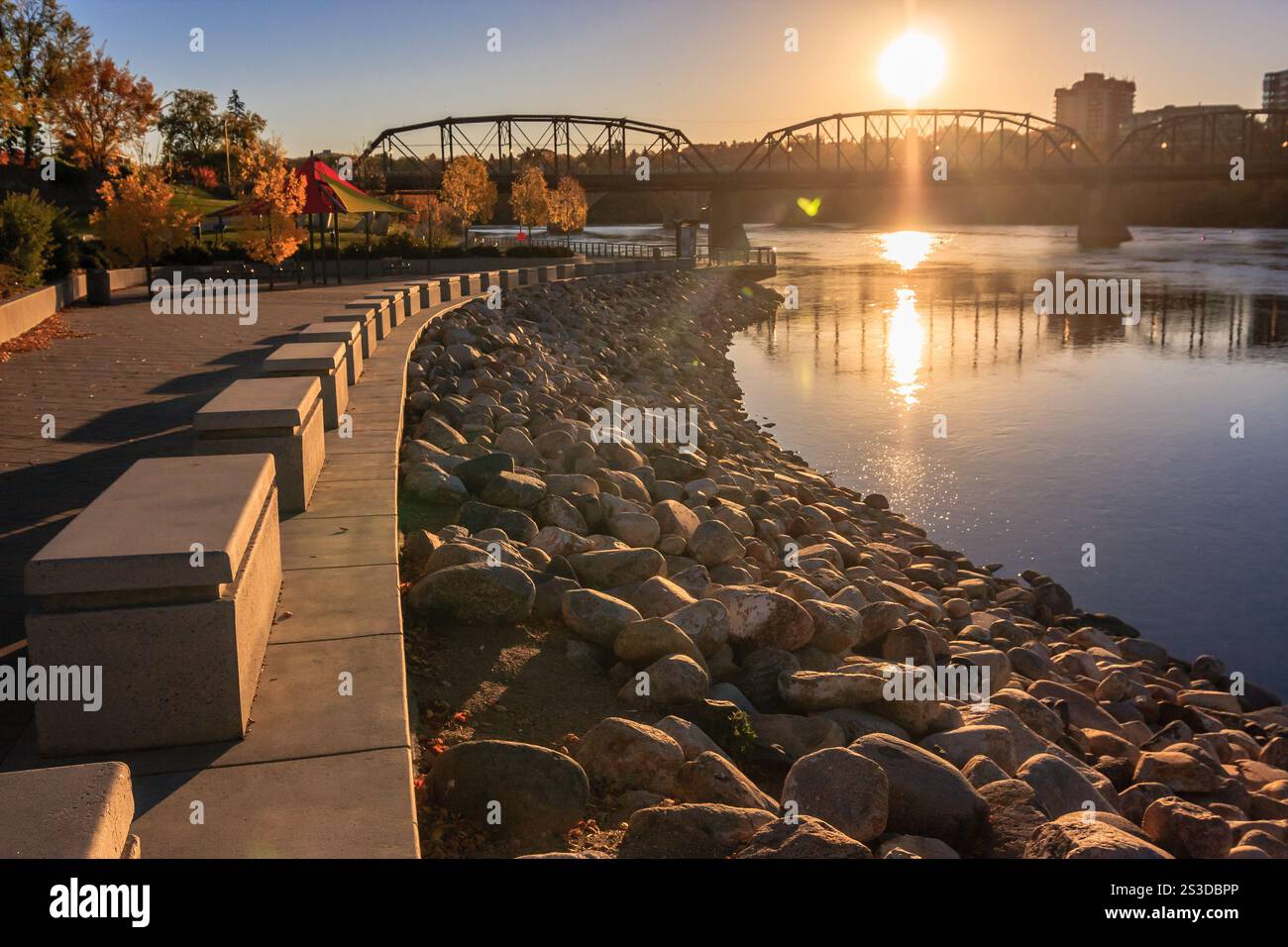 A bridge spans a river with a sunset in the background. The bridge is surrounded by a stone wall and a path Stock Photo