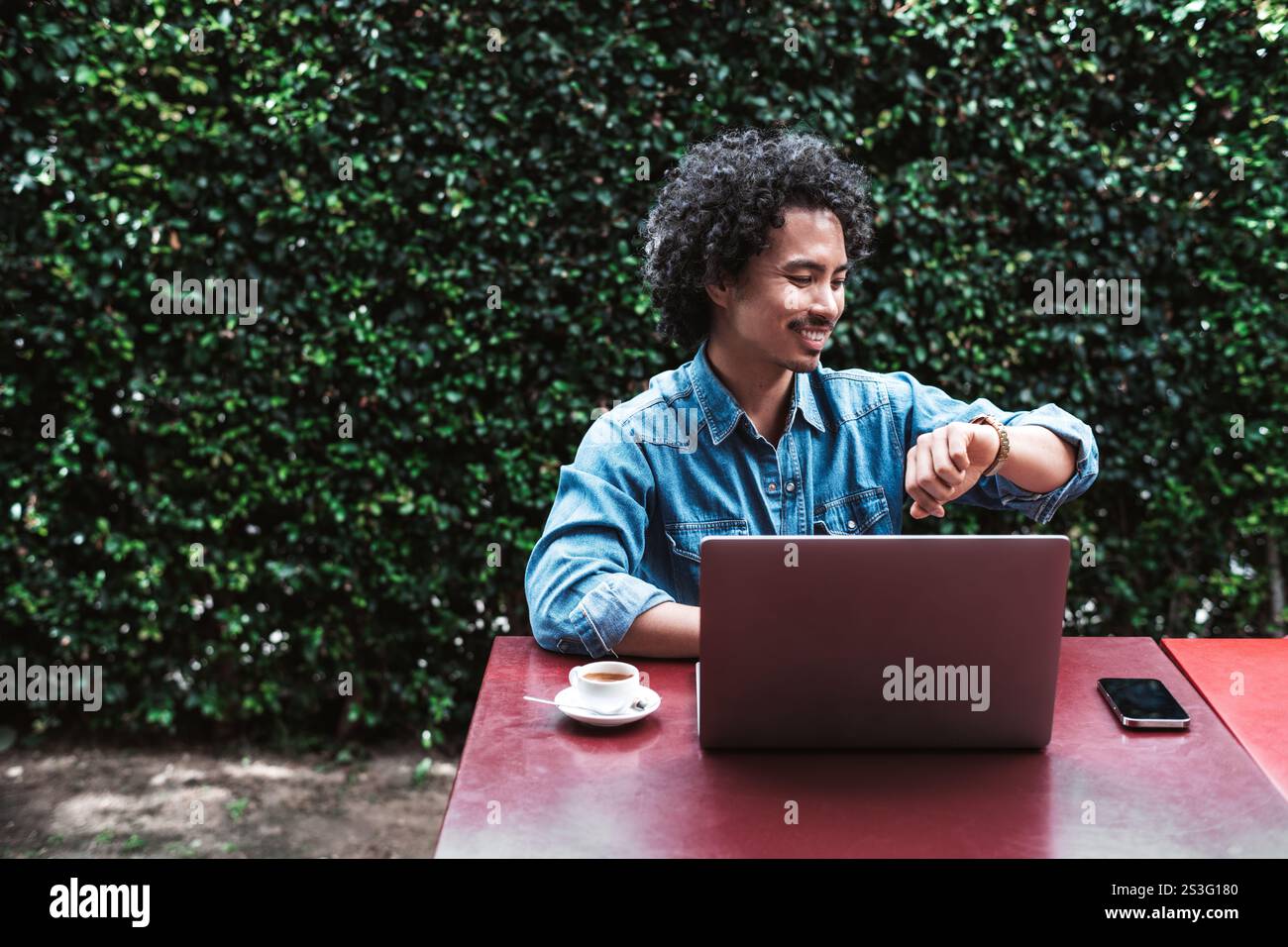 Smiling young man with curly hair sitting outdoors at a red table, working on a laptop, checking his watch, with a smartphone and coffee cup nearby, g Stock Photo