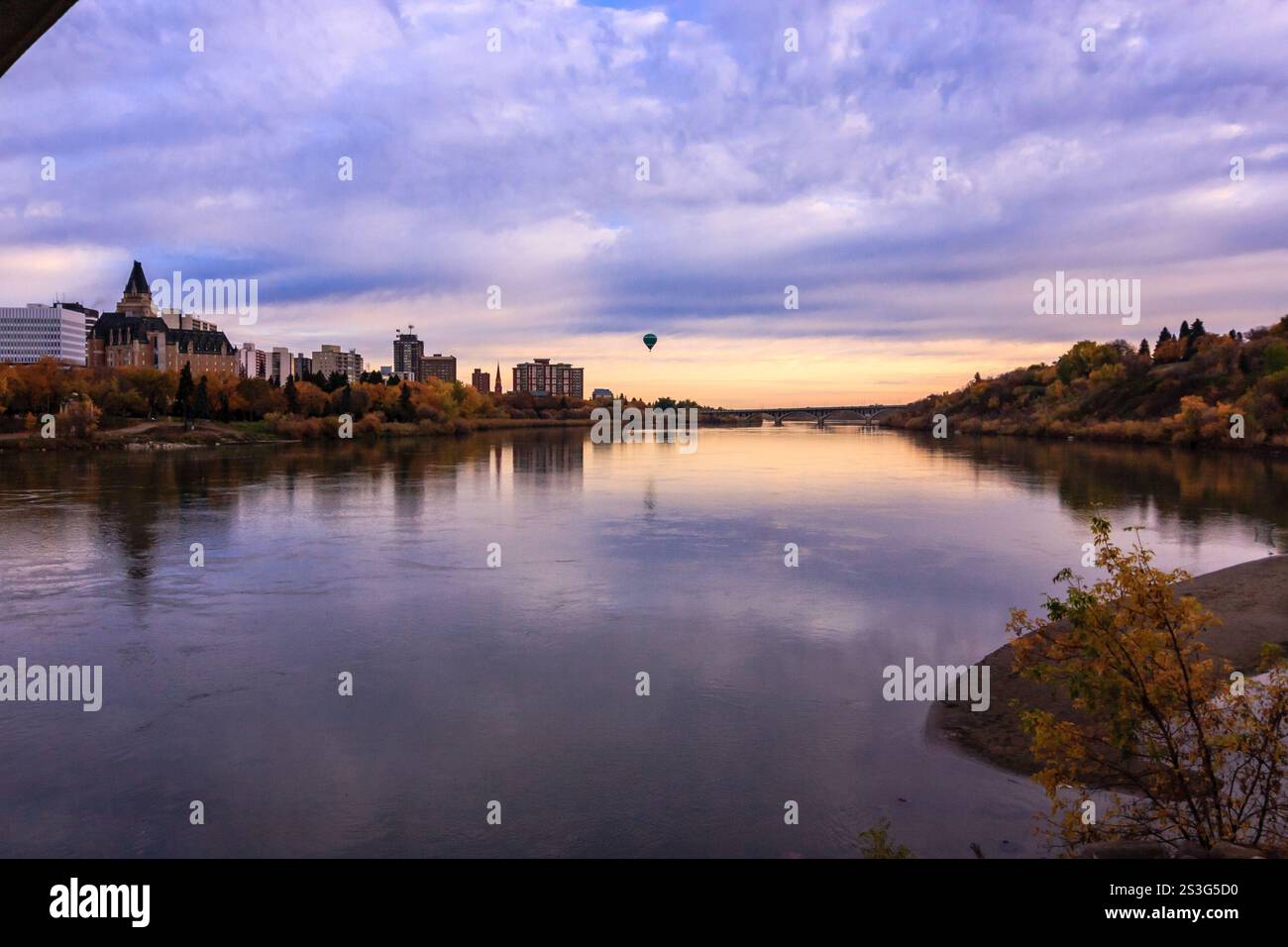 A hot air balloon is flying over a river with a city in the background. The sky is cloudy and the sun is setting Stock Photo