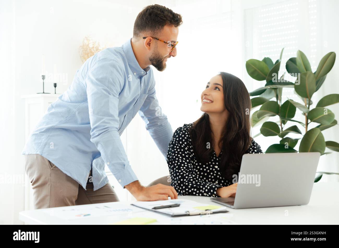Smiling arabian or indian woman in sits at a desk with a laptop, looking up at a confident man in glasses in the office, multiracial business colleagues working together on a project, discussing ideas Stock Photo