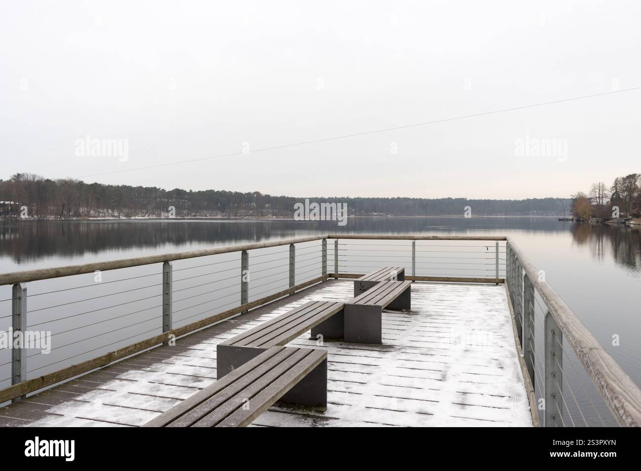 A tranquil winter scene featuring a wooden pier with snow-covered benches, overlooking a calm, frozen lake surrounded by forest. Stock Photo