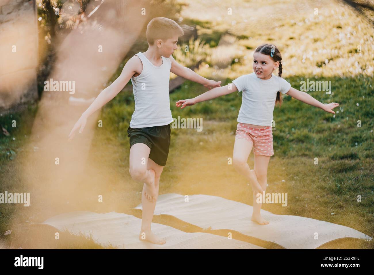 Two kids, a boy and girl, playfully balance on yoga mats in a grassy ...