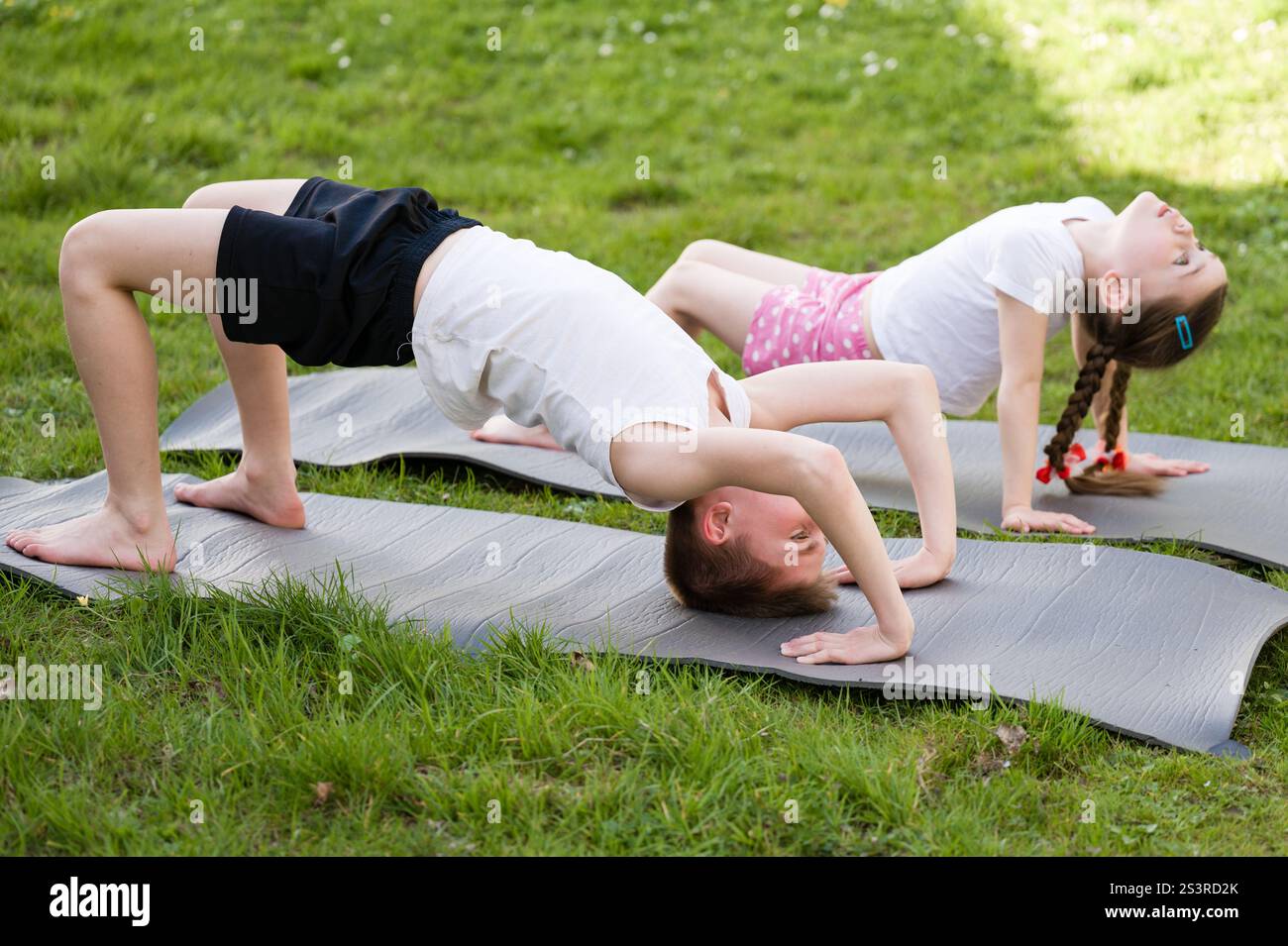 Two children practice yoga poses on mats outdoors. A boy performs a ...