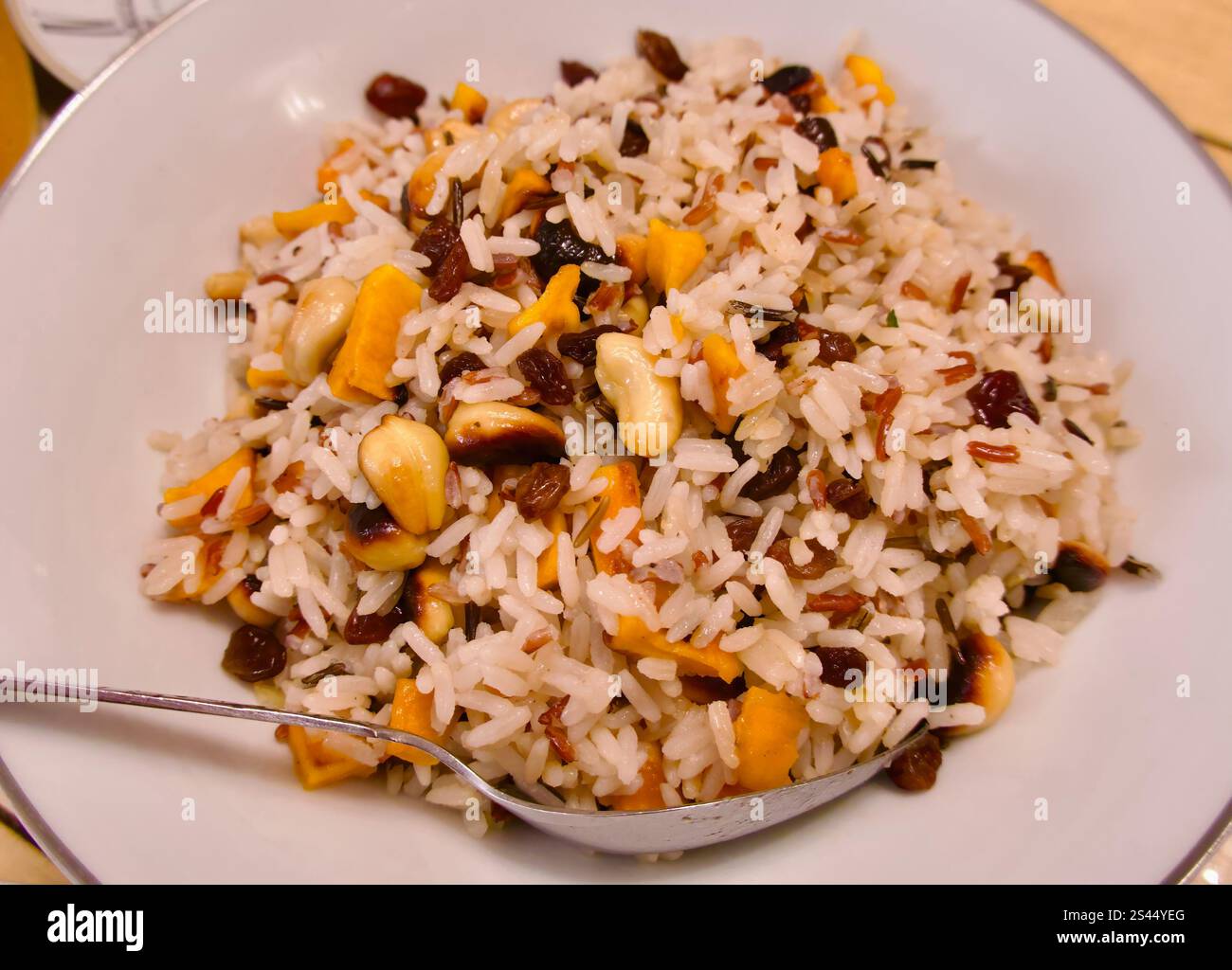 Plate of warm whole grain rice cashew nuts sultanas and pieces of dried apricot in a white dish with a serving spoon Stock Photo