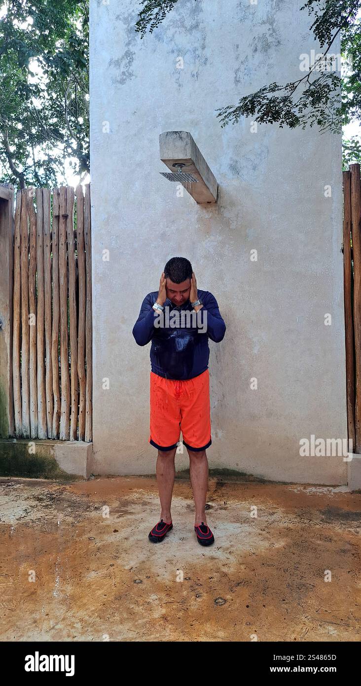 Dark-skinned Latino adult man wearing a swimsuit and aqua shoes bathes and rinses in public spa showers to remove sand, sunscreen Stock Photo