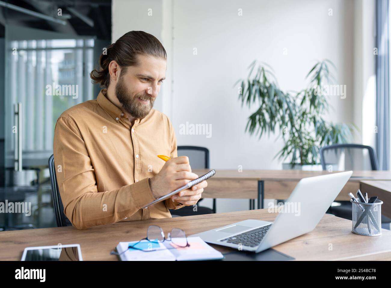 Smiling businessman working at a desk, writing in a notebook with a laptop nearby. Ideal for concepts of productivity, business planning, and professional work environments. Stock Photo