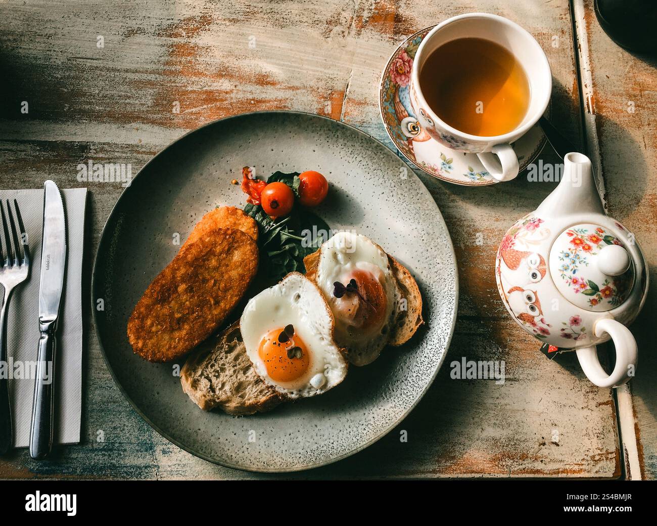 A tempting breakfast spread featuring perfectly cooked eggs on toast, crispy fried potatoes, fresh greens, and tea in an elegant teaware setup, offeri Stock Photo