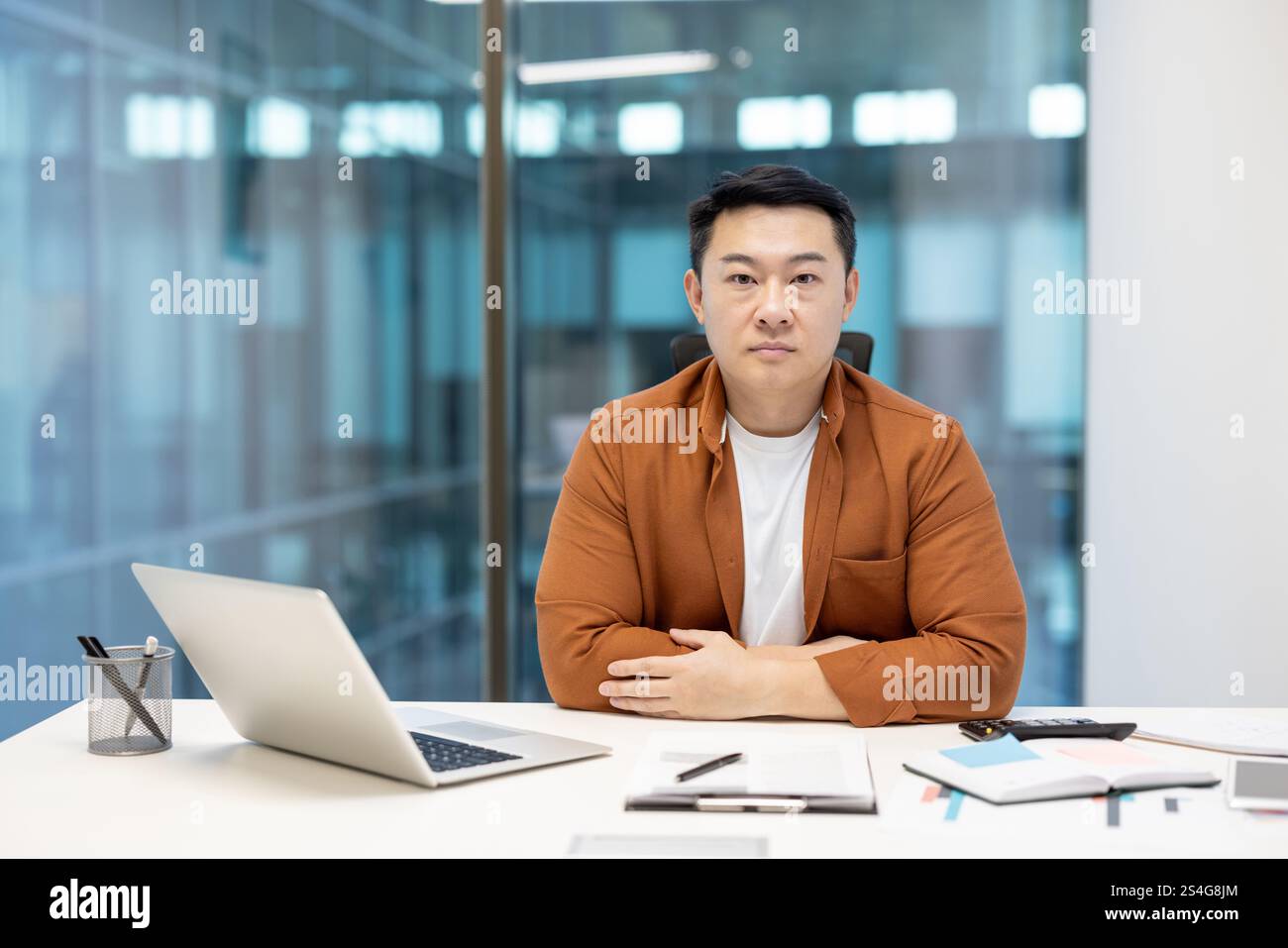 Portrait of confident serious businessman, Asian man looking focused at camera, man behind paperwork with laptop looking at camera. Stock Photo