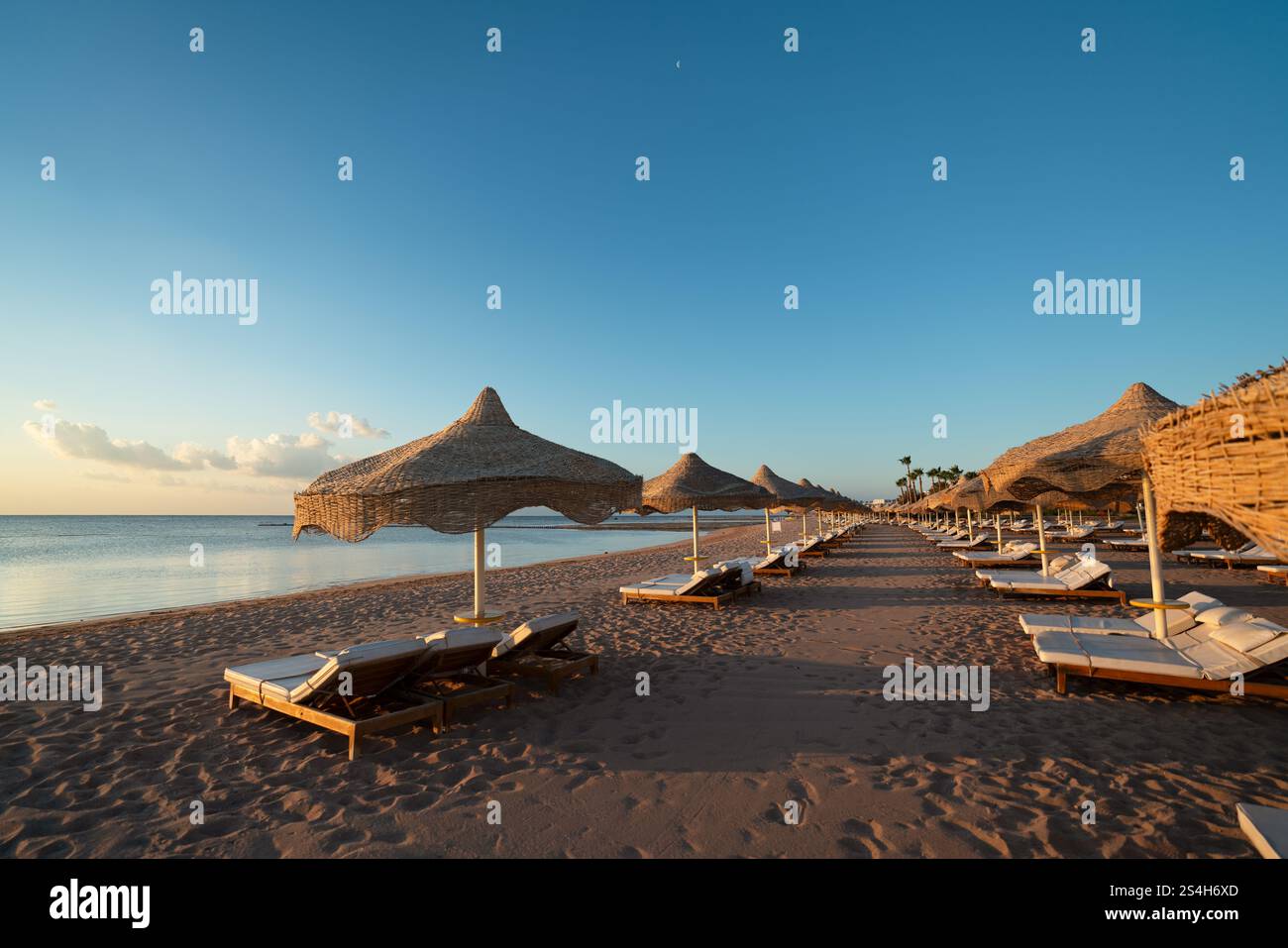 The sunrise over the sea is illuminated by sun loungers and umbrellas on a resort beach with fine sand. High resolution photo. Stock Photo