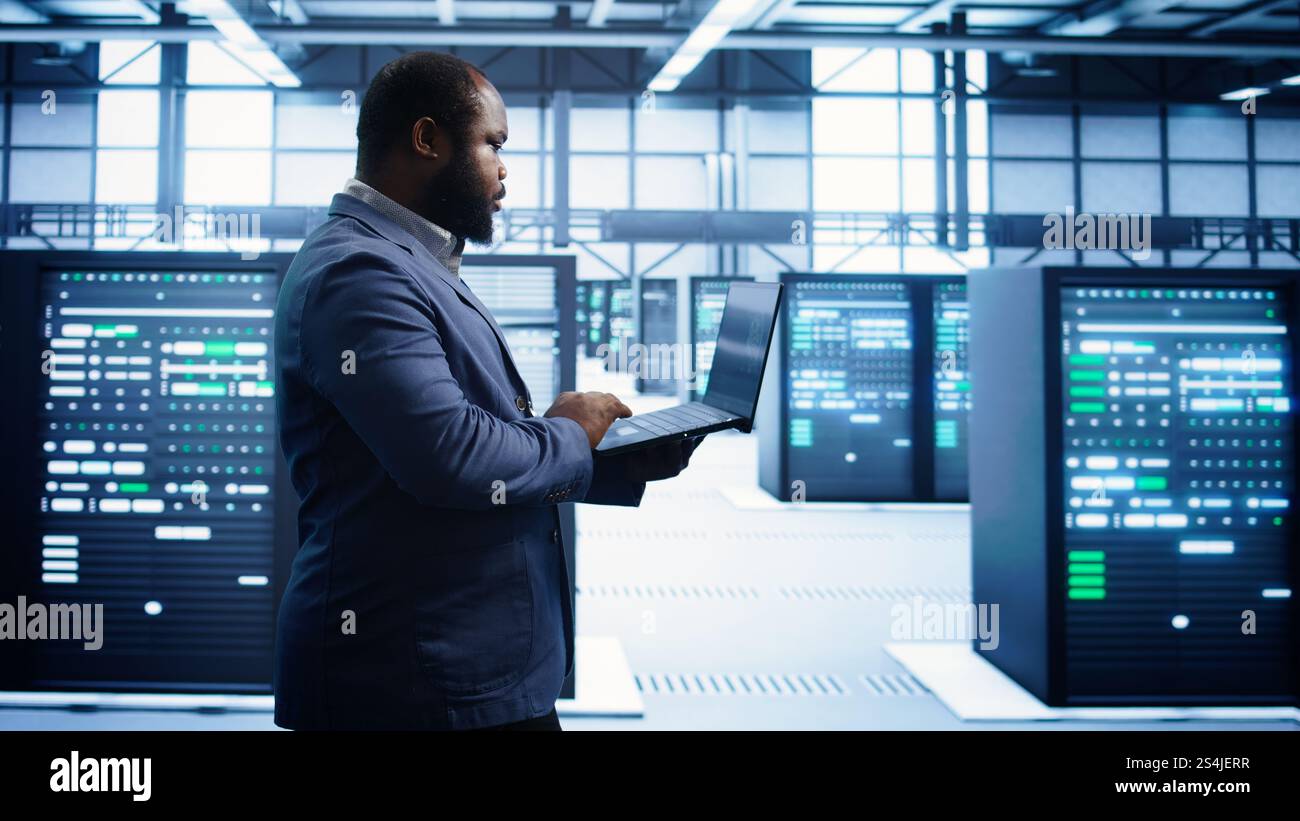 African american man walking in high tech facility, using laptop to patch network issues leading to slow data transfer rates. Data center worker fixing errors affecting supercomputers, camera B Stock Photo