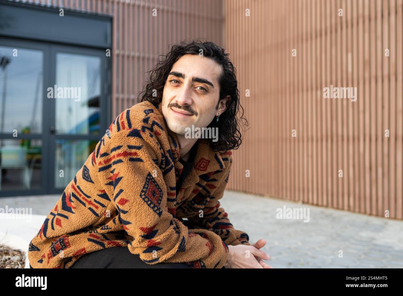 A man with long hair and a mustache, wearing a cozy patterned jacket, smiles while sitting outdoors The background shows a modern building facade, add Stock Photo