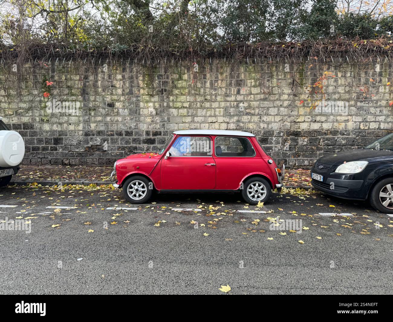 A classic red Mini Cooper parked on a street between two other cars, with a stone wall in the background and fallen leaves on the ground. - Smartphone Captured Stock Image