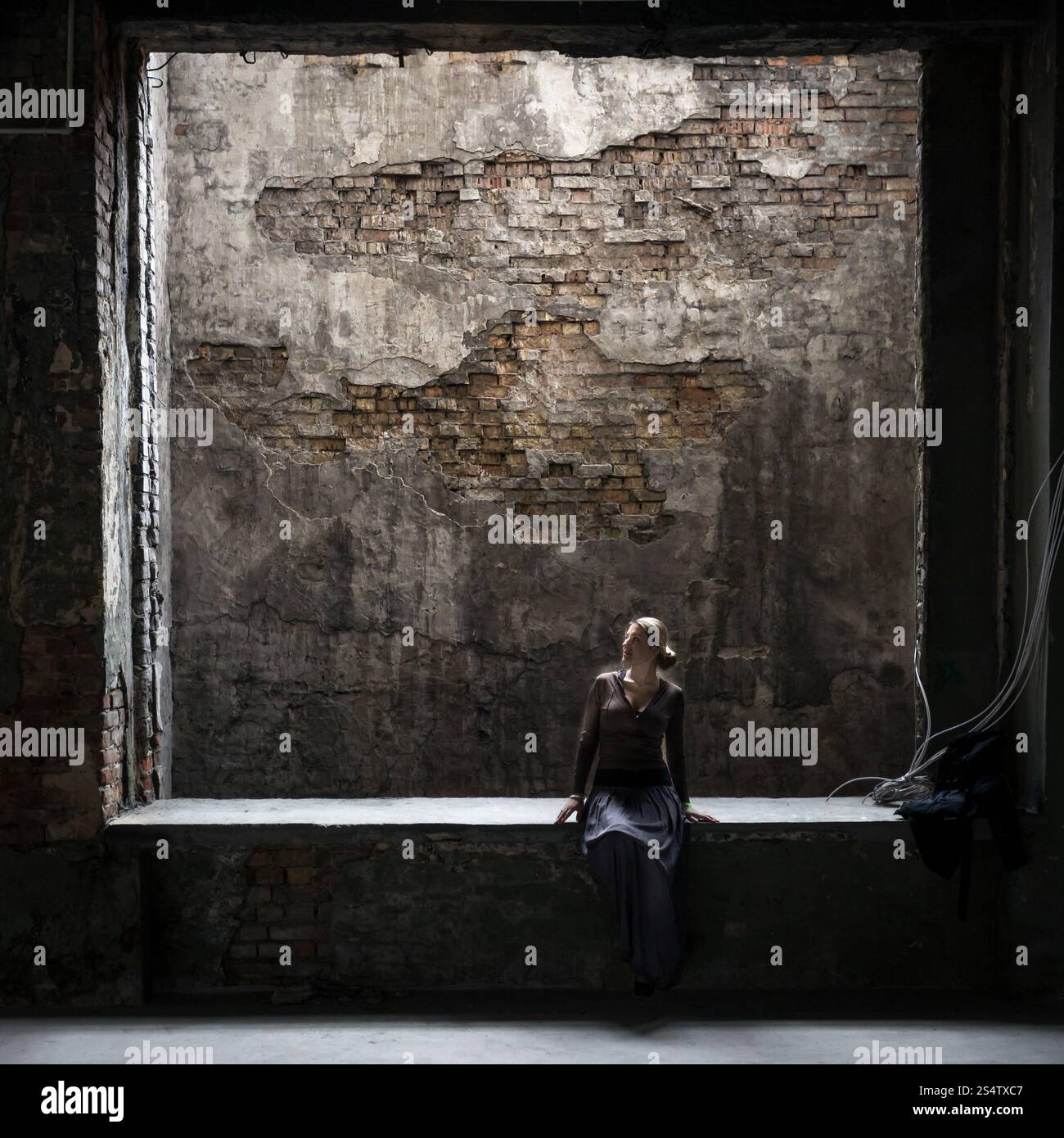 Grungy photo of lonely woman sitting at big window in old abandoned building Stock Photo