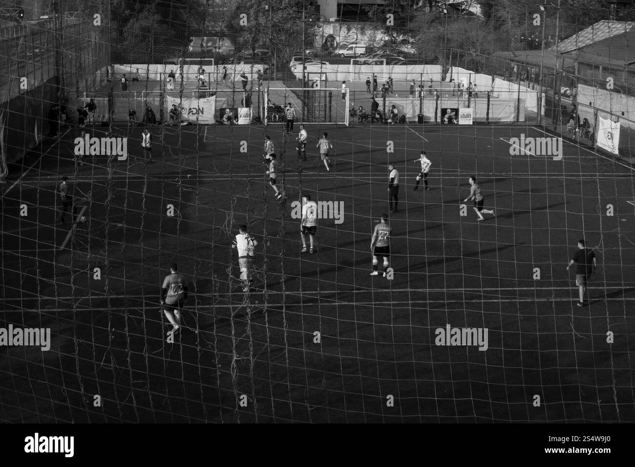 Black-and-white photo of an outdoor soccer match on an urban field, viewed through netting. Captures community, teamwork, and local sports atmosphere. Stock Photo