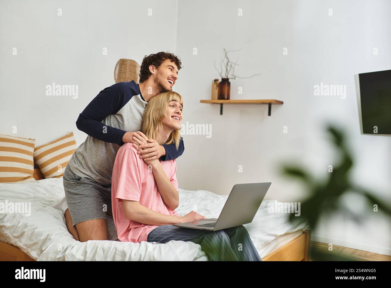 Two men share a loving embrace while using a laptop in a comfortable bedroom setting. Stock Photo