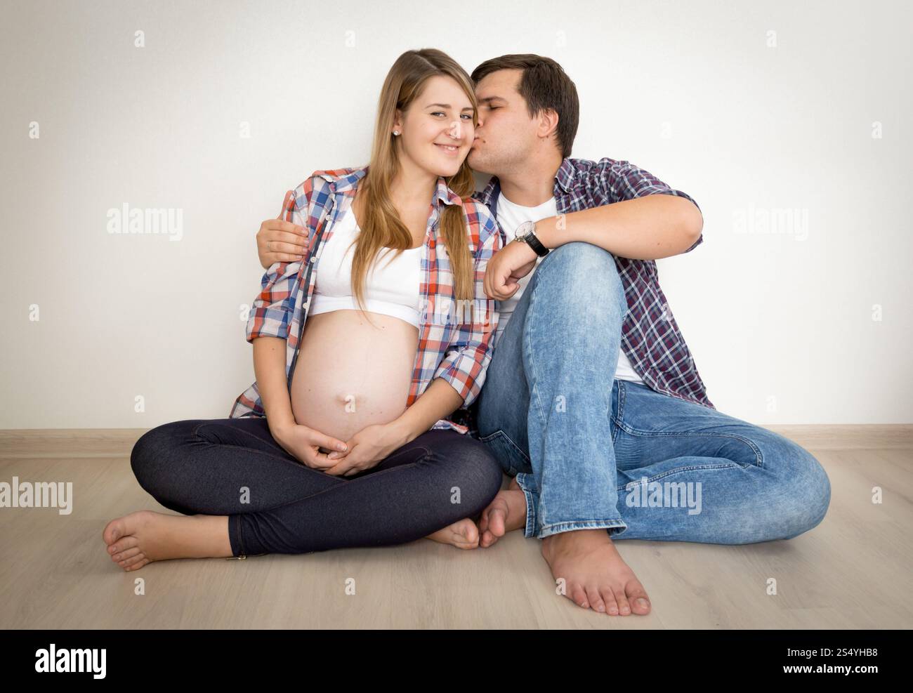 Toned portrait of happy pregnant couple sitting on floor and kissing Stock Photo