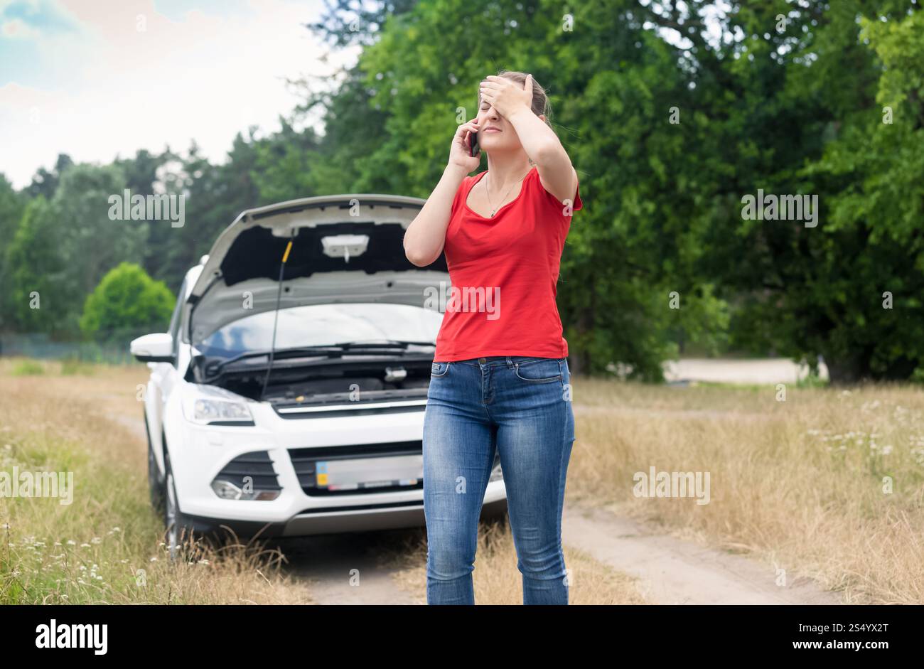 Sad woman standing at broken car and calling on cellphone for help Stock Photo